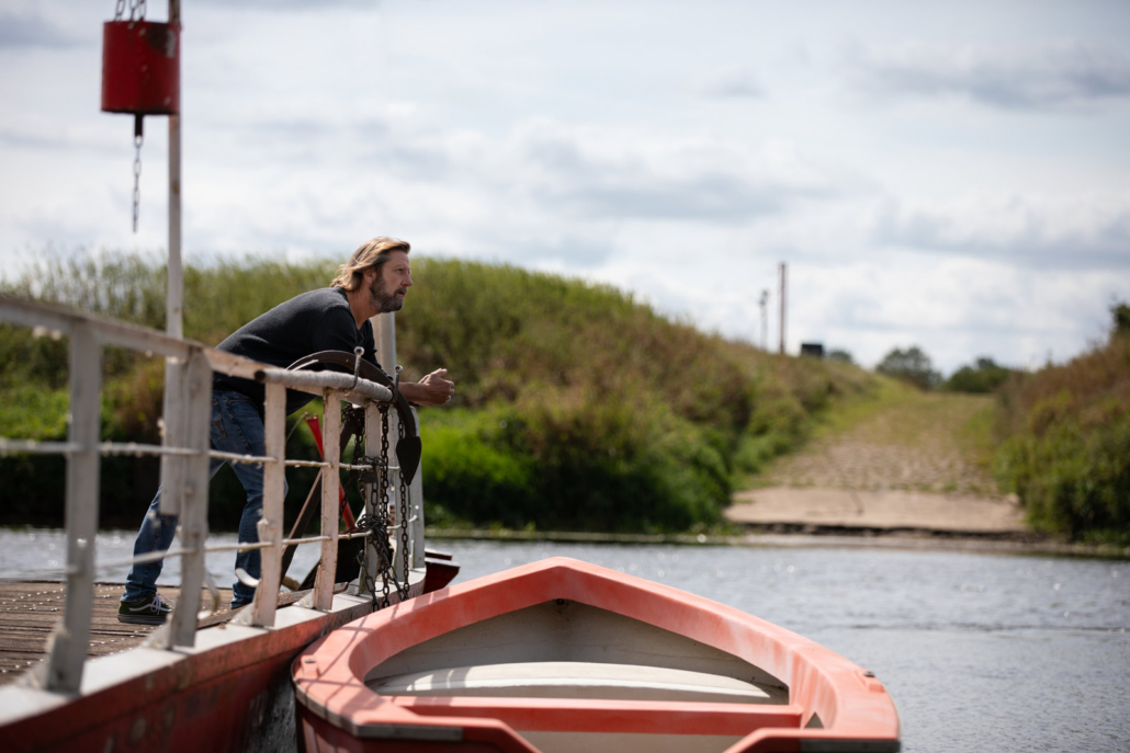 Frank Botje steht an der Reling am Fluss. Vor ihm ein Boot, auf der anderen Uferseite führt ein Weg weiter.